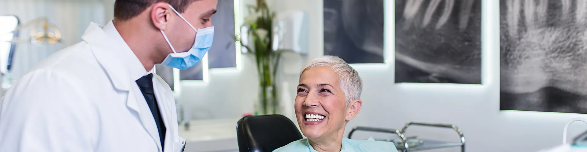 Doctor with clipboard in mask looking at smiling patient in dental chair.
